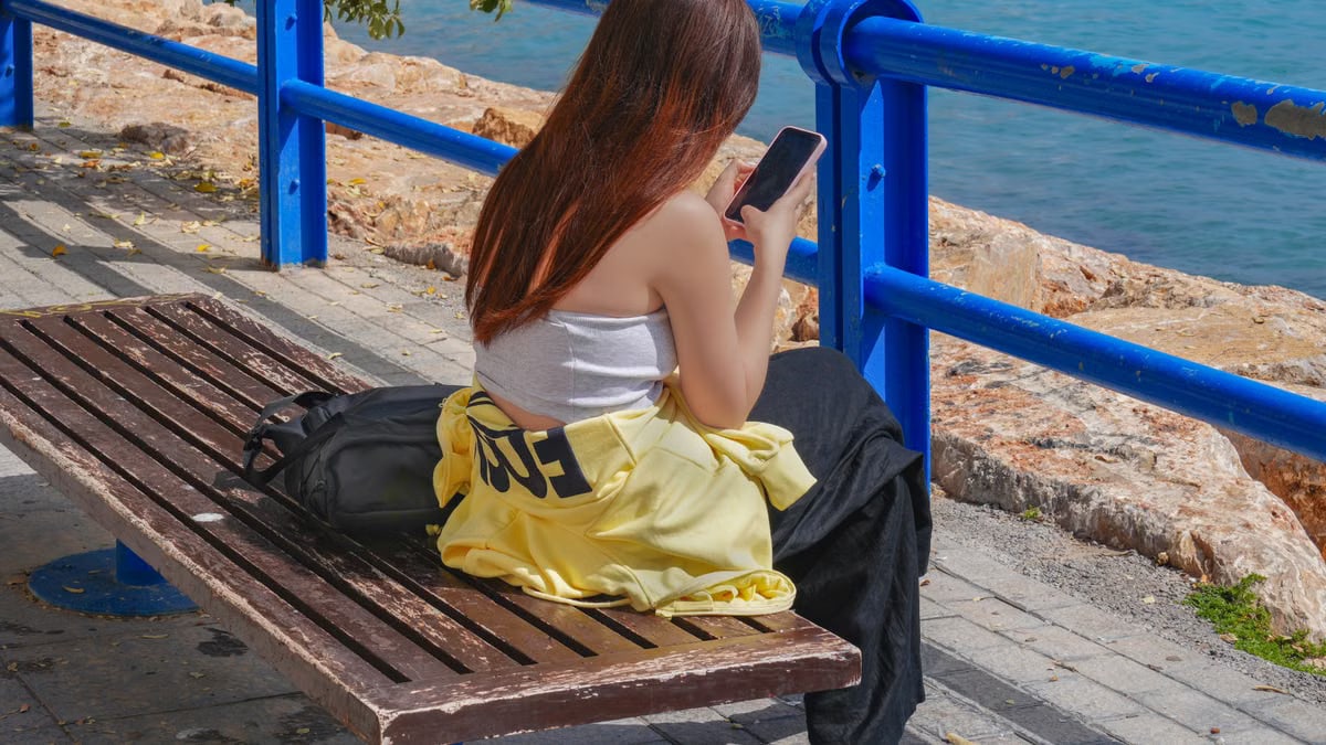 A young woman sits on a bench using her smartphone by the seaside in Alicante, Spain.